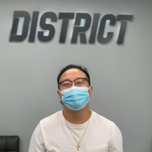 A man with a blue medical mask on standing infront of a wall with a sign that reads District.