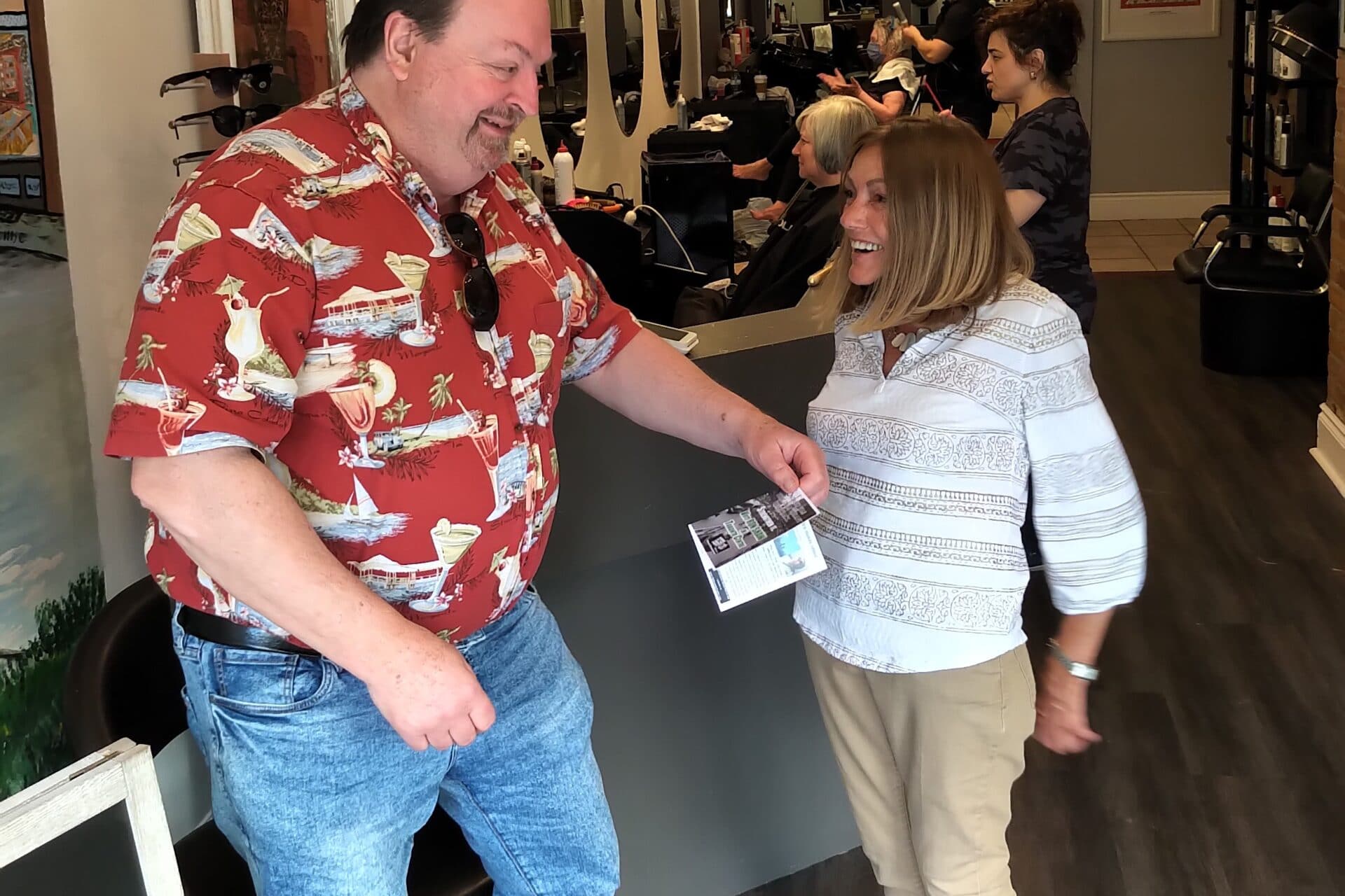 A man in a red shirt and blue jeans gives a flyer to a persn wearing a white and grey shirt and beige pants.