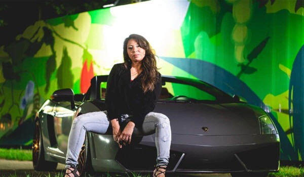 Trini Nish, the star of ERIKA: Fringe Edition, leans again a car against a green background