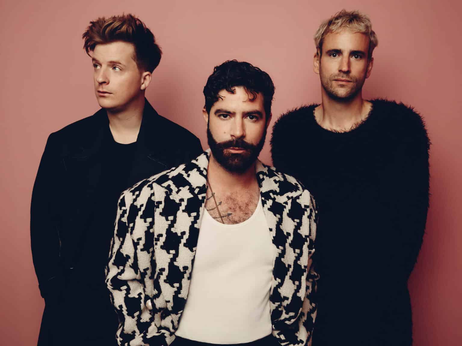 The three members of Foals are wearing black and white, standing in front of a brown wall