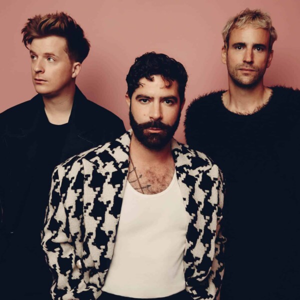 The three members of Foals are wearing black and white, standing in front of a brown wall