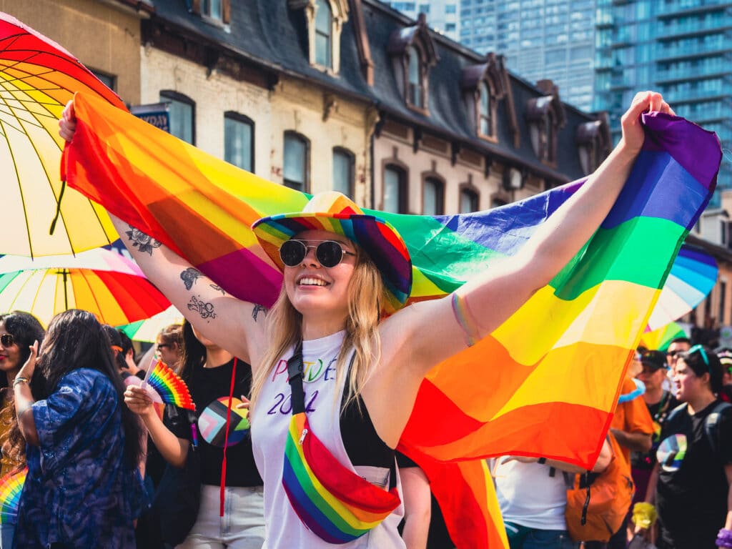 A person with long blonde hair, a rainbow cowboy hat and a rainbow fanny pack, holding a rainbow flag at the pride parade.