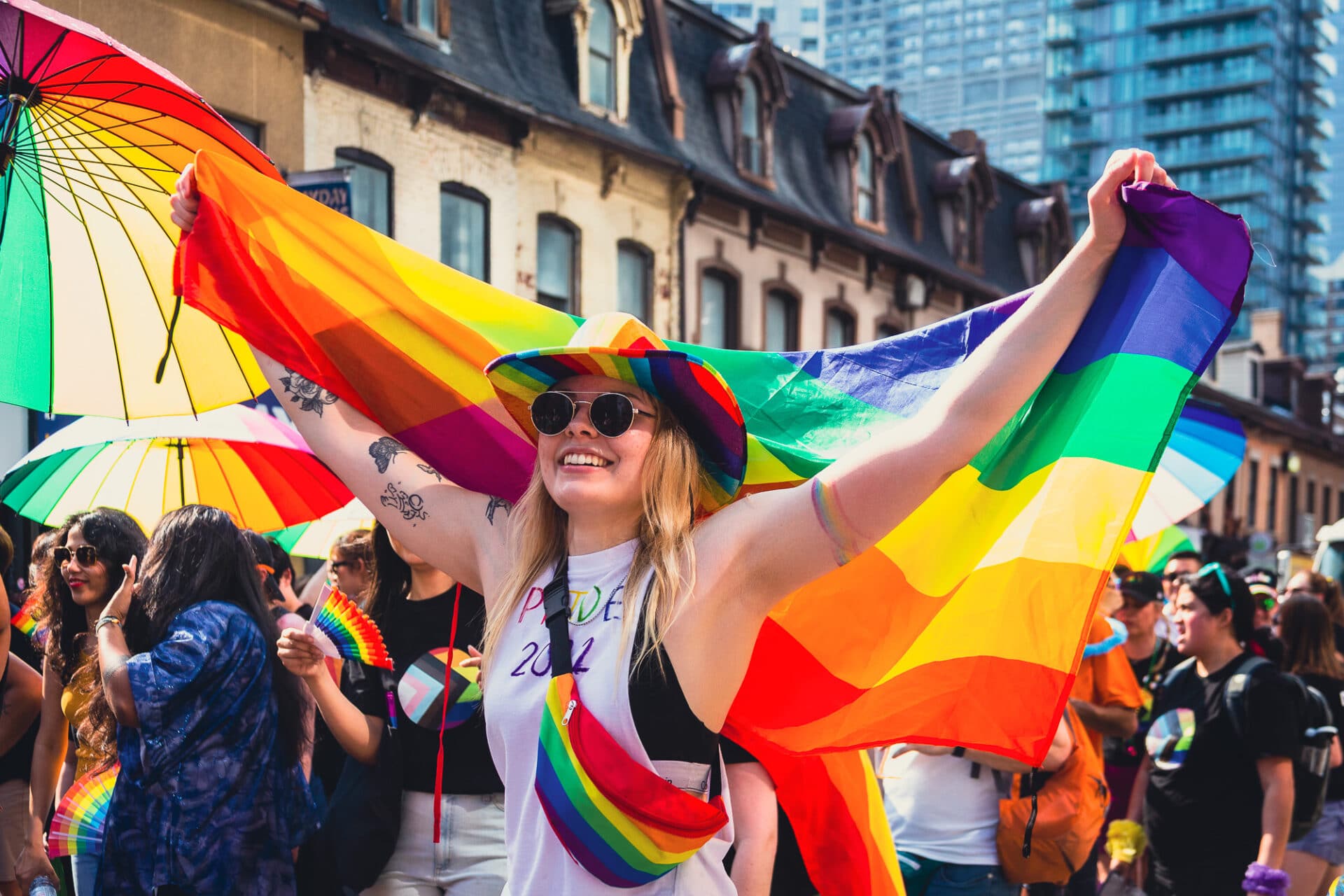 A person with long blonde hair, a rainbow cowboy hat and a rainbow fanny pack, holding a rainbow flag at the pride parade.