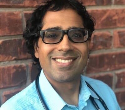 A person with a stethoscope, light blue colloared shirt and black framed glasses smiles against a brick wall background.