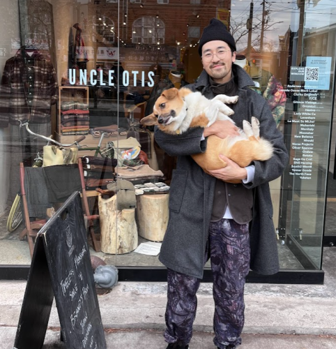 A person with a grey clothing is holding a dog as he stands in front of a glass storefront.